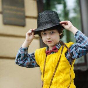 A stylish young boy wearing a yellow sweater, plaid shirt, and black hat poses playfully outdoors.